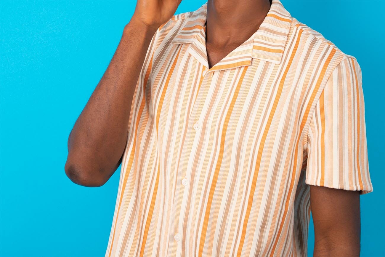 young man wearing Hawaiian striped shirt over blue background laughs happily keeps hand on chin expresses positive emotions smiles broadly has carefree expression