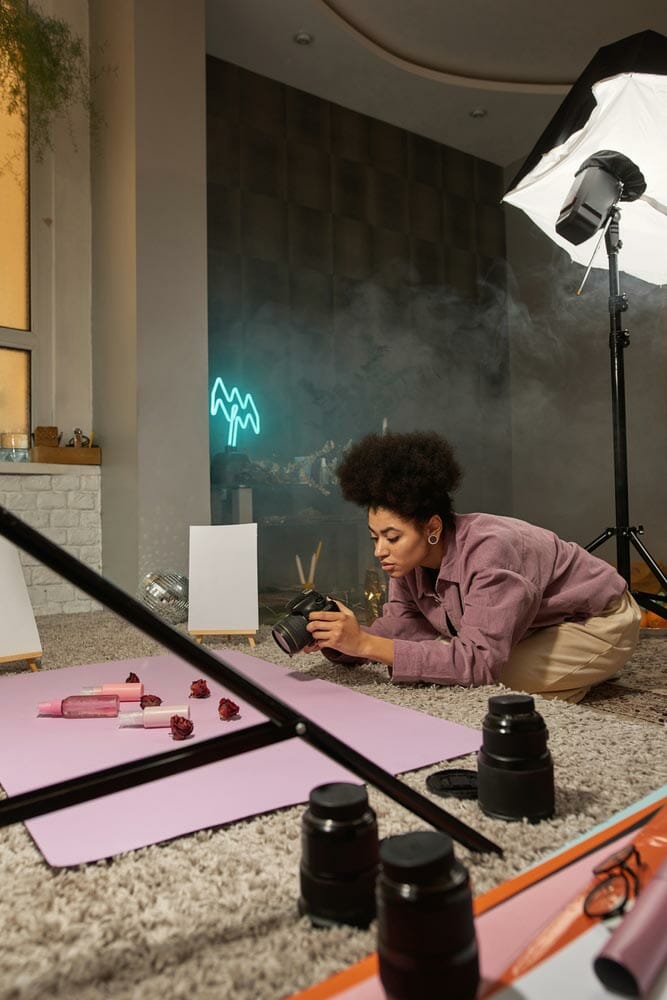 Black female photographer taking photo of bottles with cosmetic cream and lotion, dry rose flowers on digital camera at studio. Beauty and body care content for photostocks, commerce and advertising